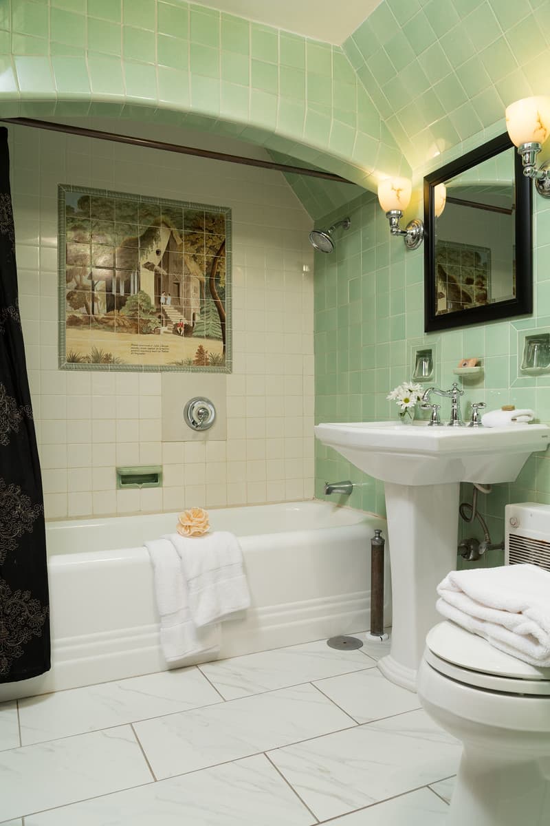 A vintage-style bathroom featuring green tiled walls, a bathtub with a towel, a pedestal sink, and a decorative wall mural.