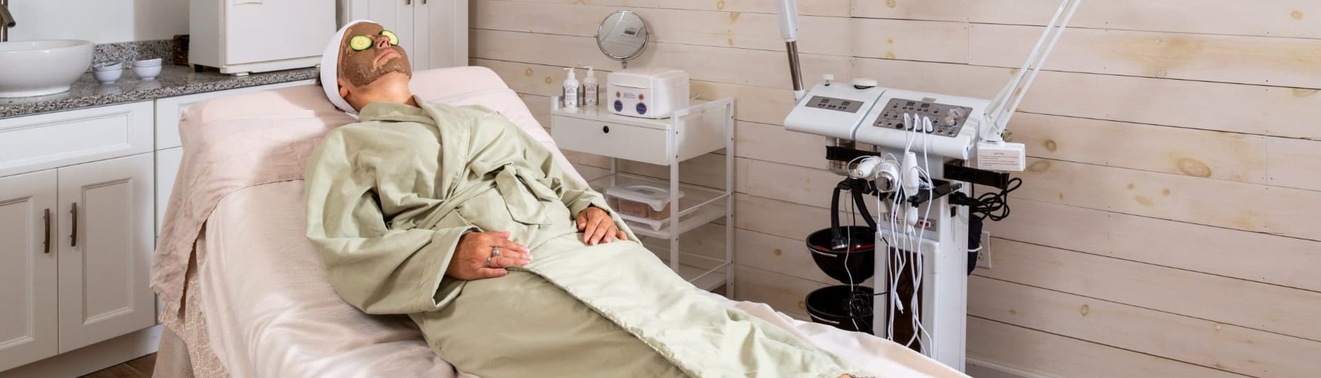 A woman in a spa robe is lying on a treatment table, wearing a mud mask and cucumber eye patches. A spa machine is visible in the background.