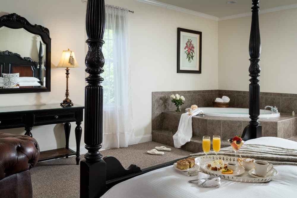 A luxurious hotel room featuring a four-poster bed, a freestanding bathtub, a breakfast tray on the bed, and a vanity table with a lamp.