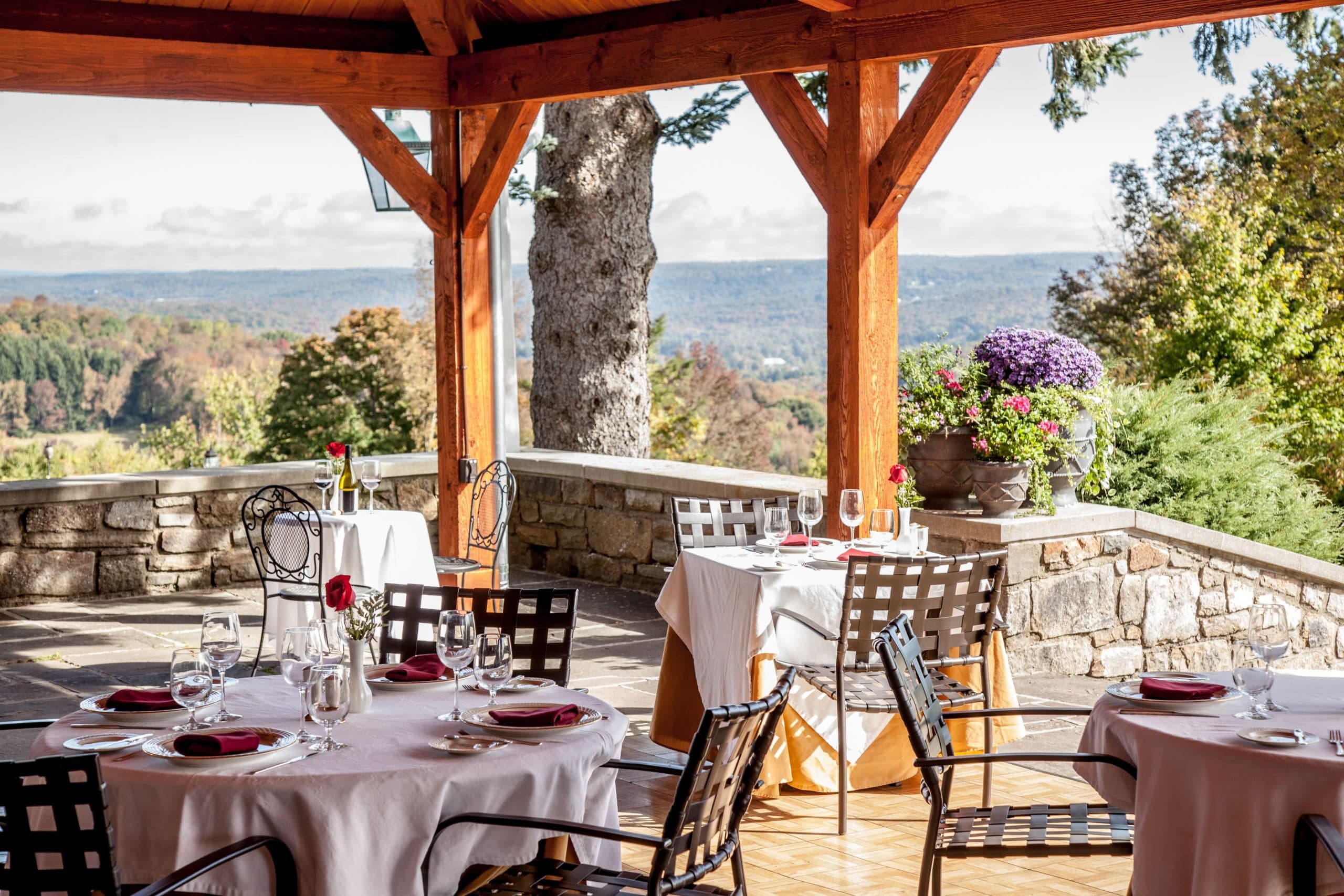 An outdoor dining area with a covered pavilion and stone walls. Tables with white tablecloths and chairs are set up on the patio, and there is a stunning view of the surrounding mountains and fall foliage.