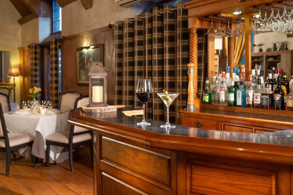 A cozy bar area with a wooden bar counter and plaid curtains. A glass of red wine and a martini glass are sitting on the counter, and a well-stocked bar is visible behind it. In the background, there is a dining area with tables and chairs.