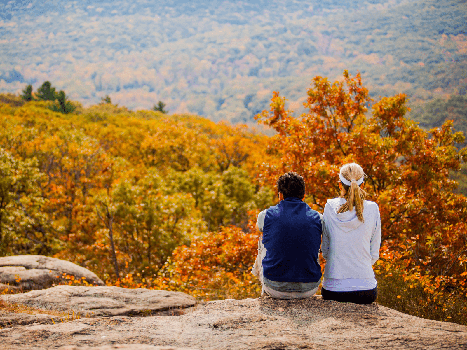 A couple sits on a rock, overlooking a vibrant autumn landscape.