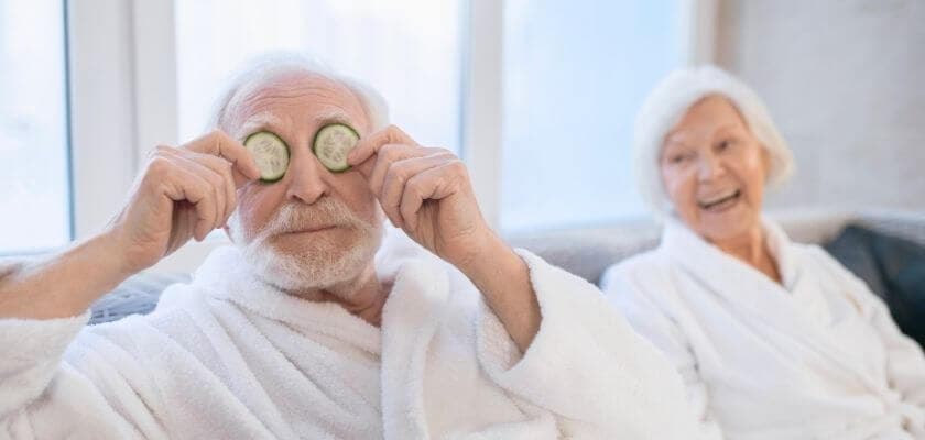 An elderly couple relaxes in bathrobes, with the man playfully holding cucumber slices over his eyes while the woman laughs.
