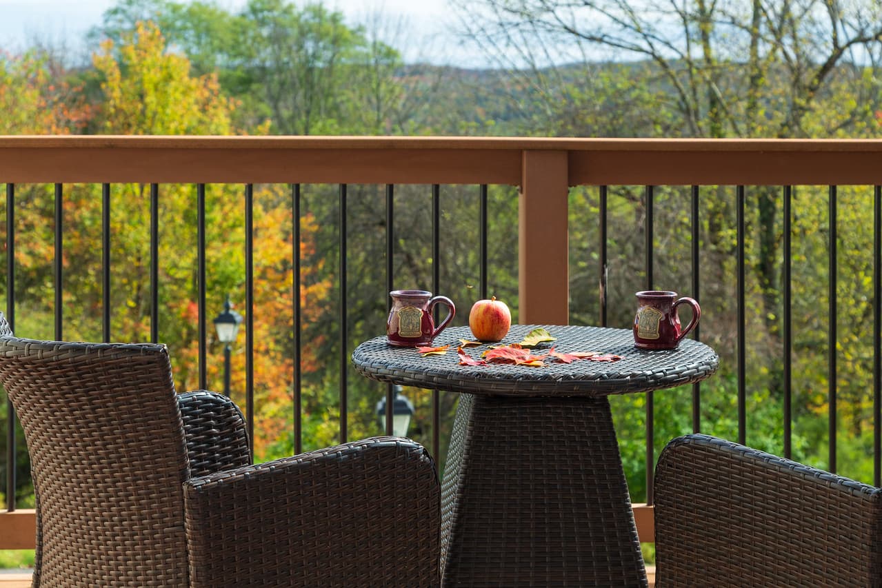 A cozy outdoor patio features a round table with two mugs and an apple, surrounded by autumn foliage.