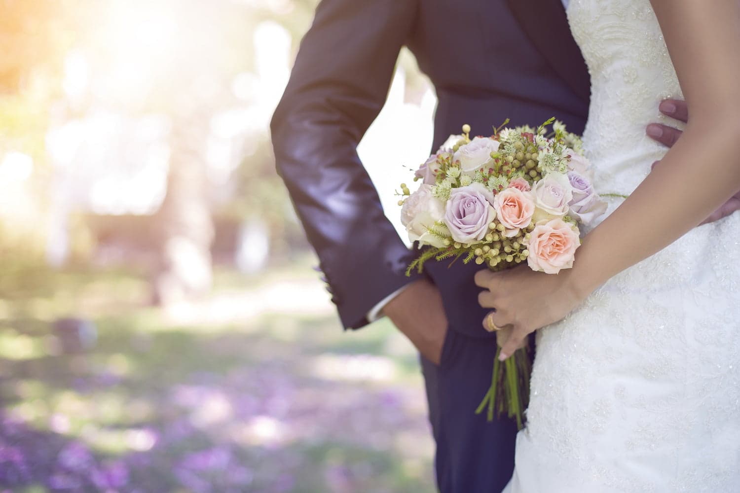A bride in a white dress holds a floral bouquet while standing next to a groom in a navy suit, set in a softly lit outdoor setting. A bride in a white dress holds a floral bouquet while standing next to a groom in a navy suit, set in a softly lit outdoor setting.