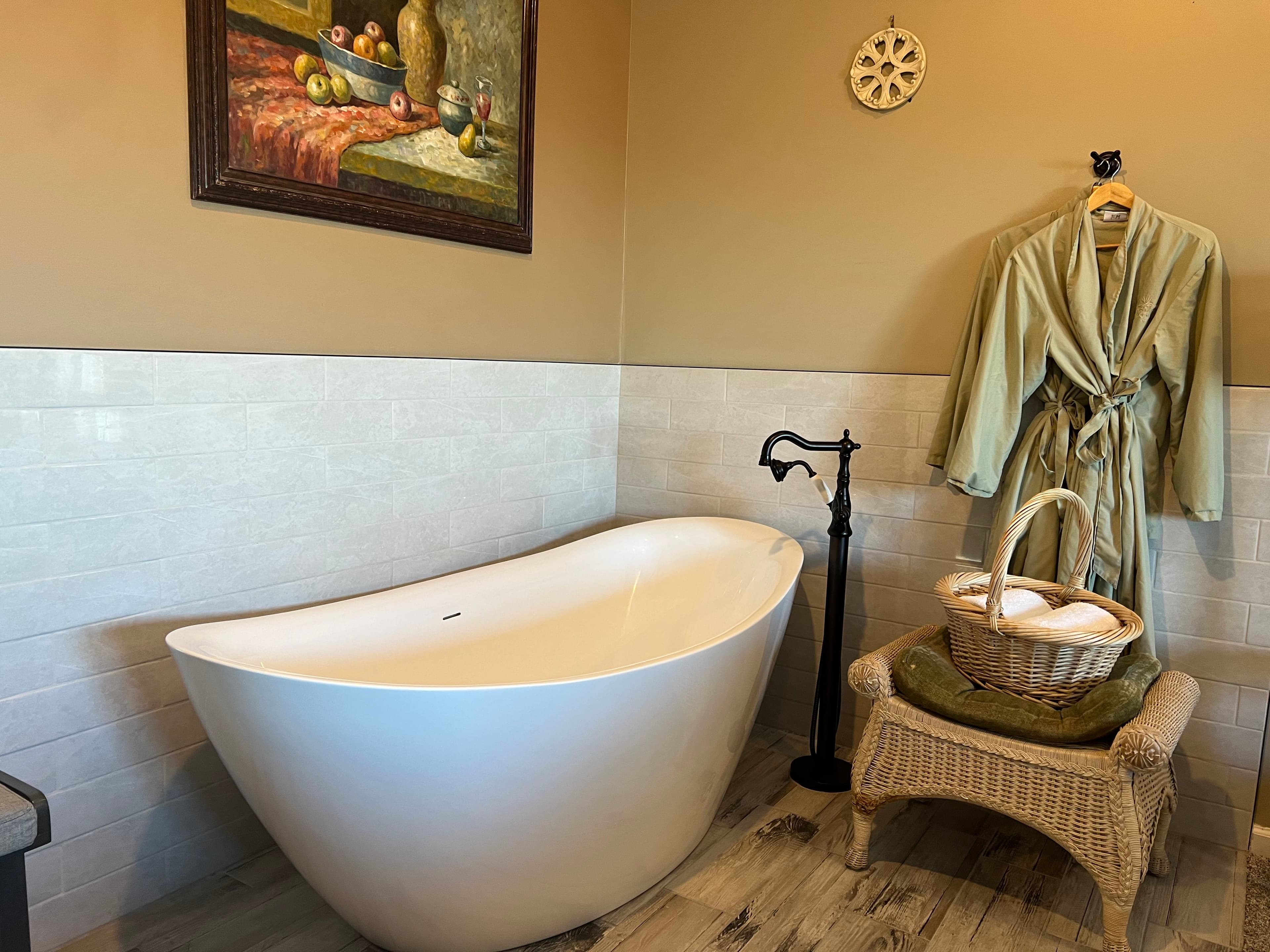 A modern bathroom featuring a freestanding white bathtub, a green robe hanging on a hook, and a wicker basket with towels.