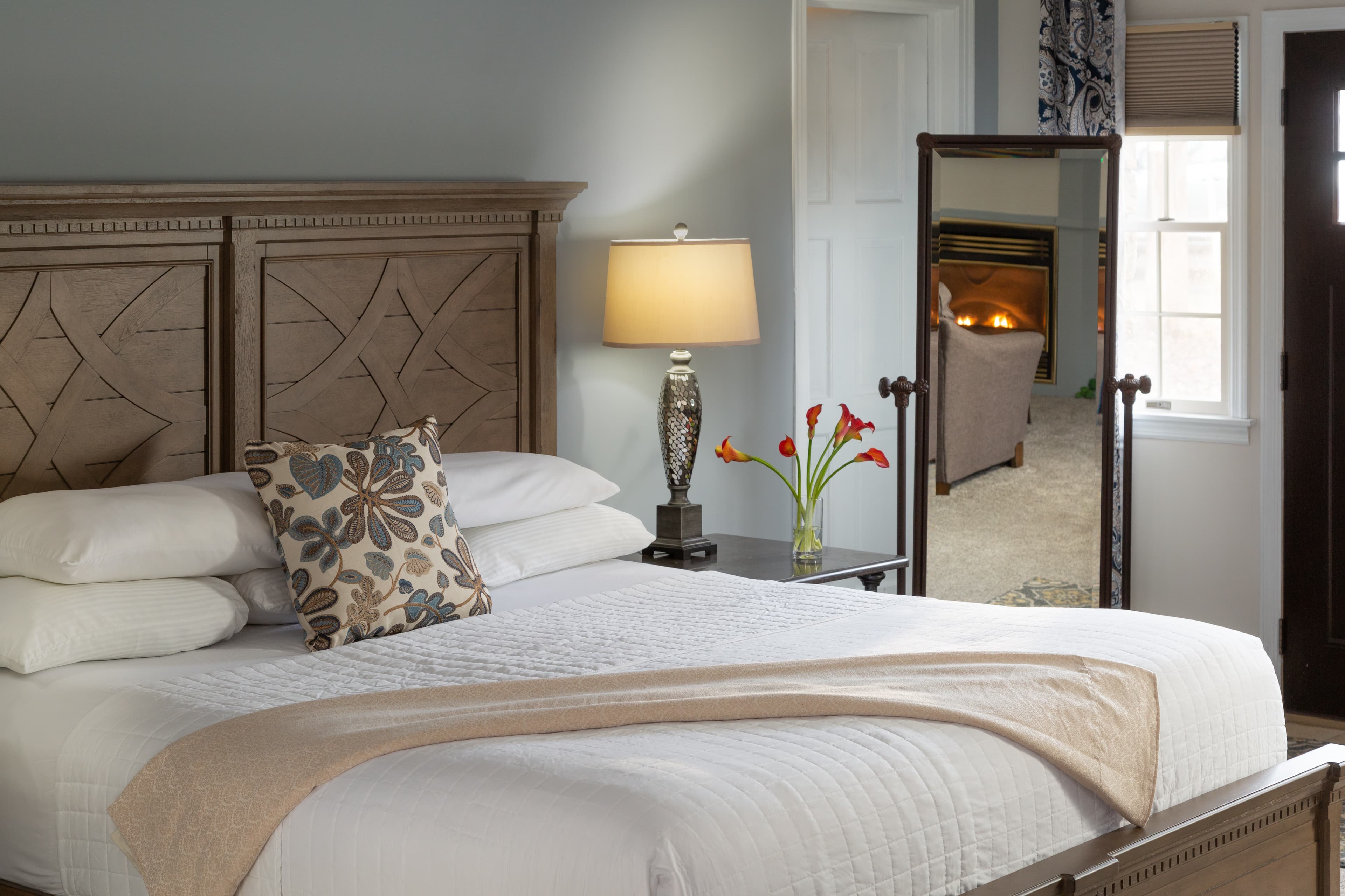 A cozy bedroom featuring a wooden headboard, a neatly made bed with decorative pillows, a lamp, and a vase of flowers.