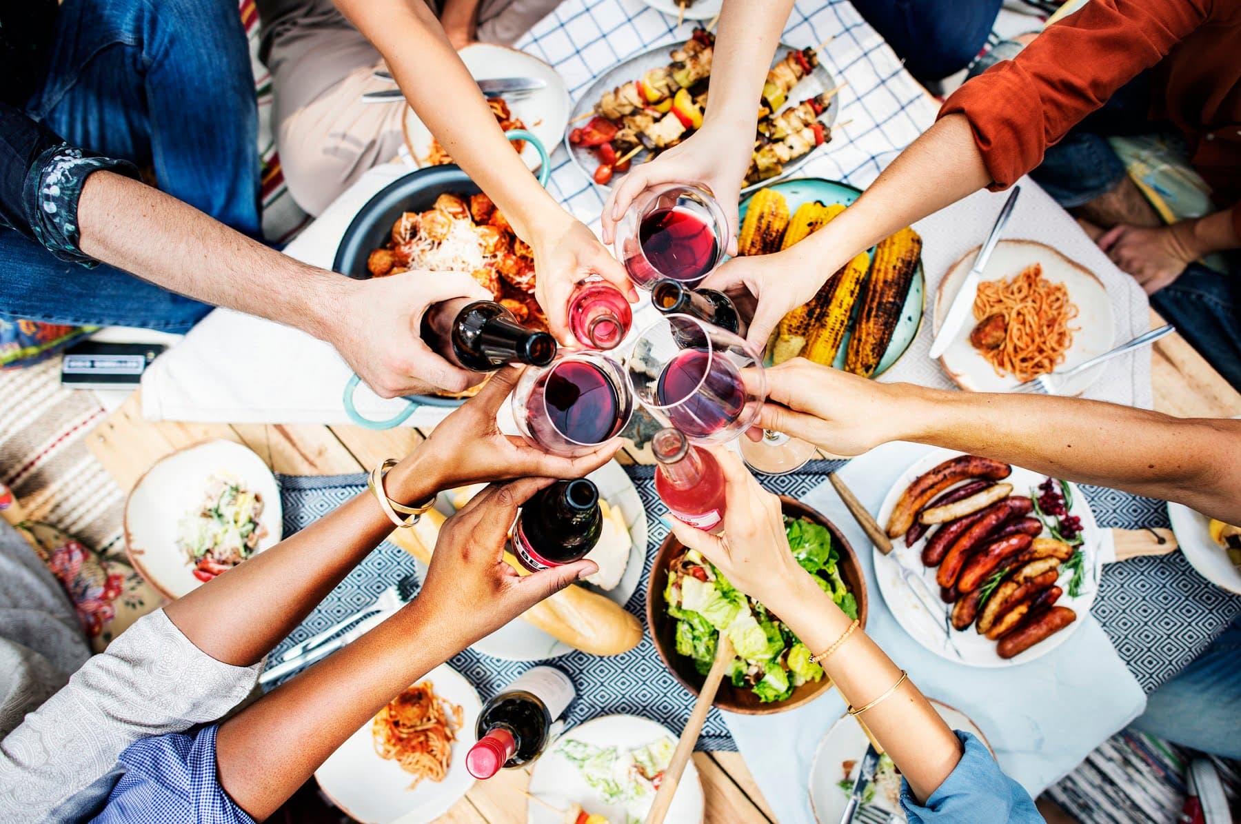 A group of hands toasting with glasses of wine above a picnic spread of various foods.