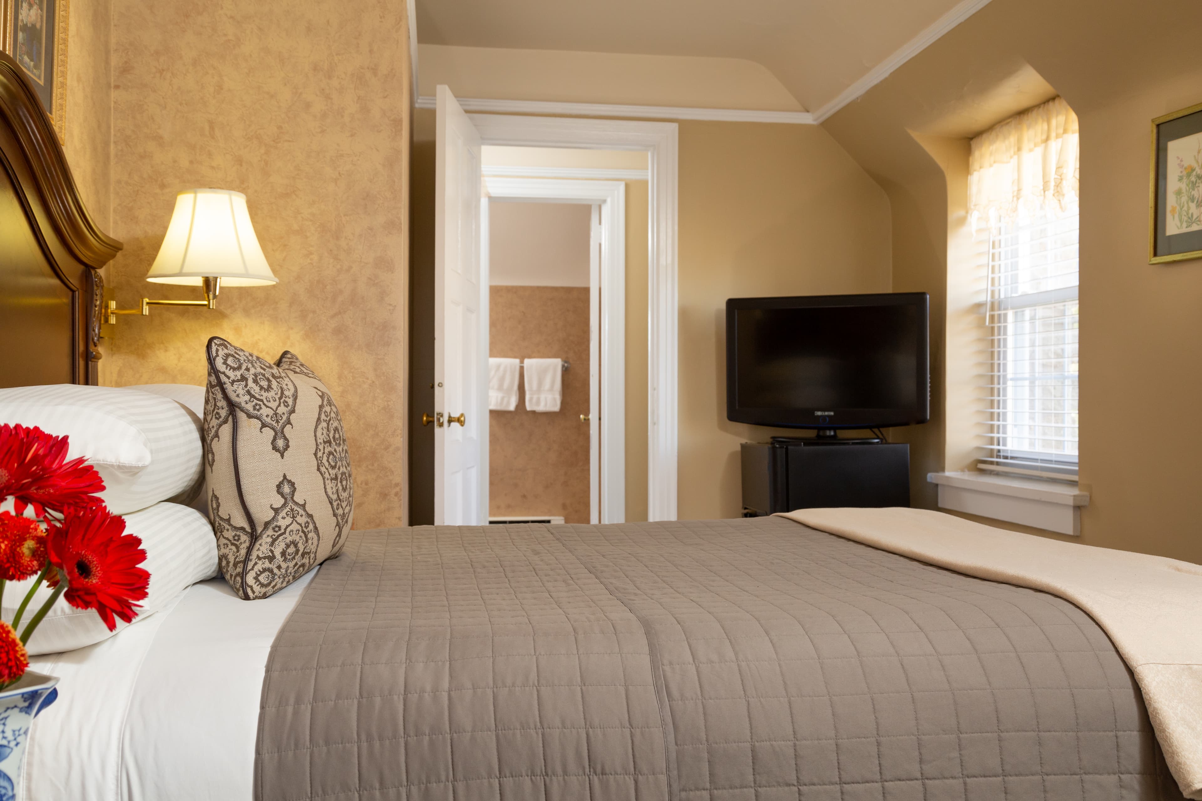 A cozy bedroom featuring a neatly made bed, decorative pillows, a television, and an open doorway leading to a bathroom.