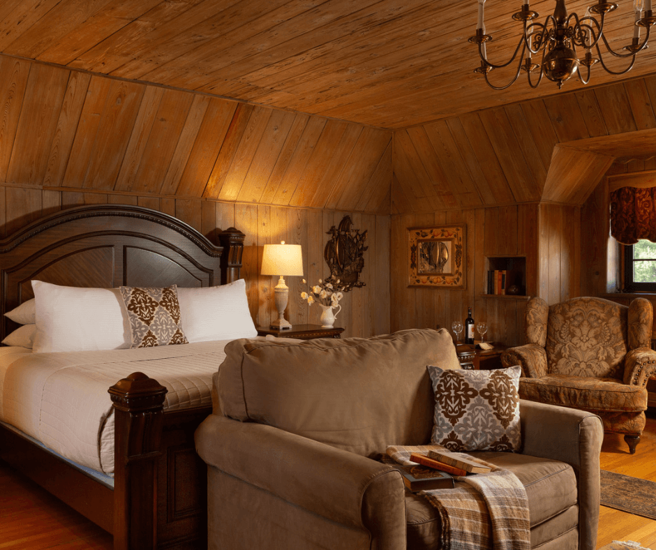 A cozy wooden-paneled bedroom featuring a large bed, an armchair, and a chandelier.
