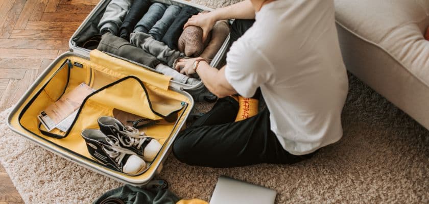 A person sits on the floor packing a suitcase with neatly rolled clothes and shoes.