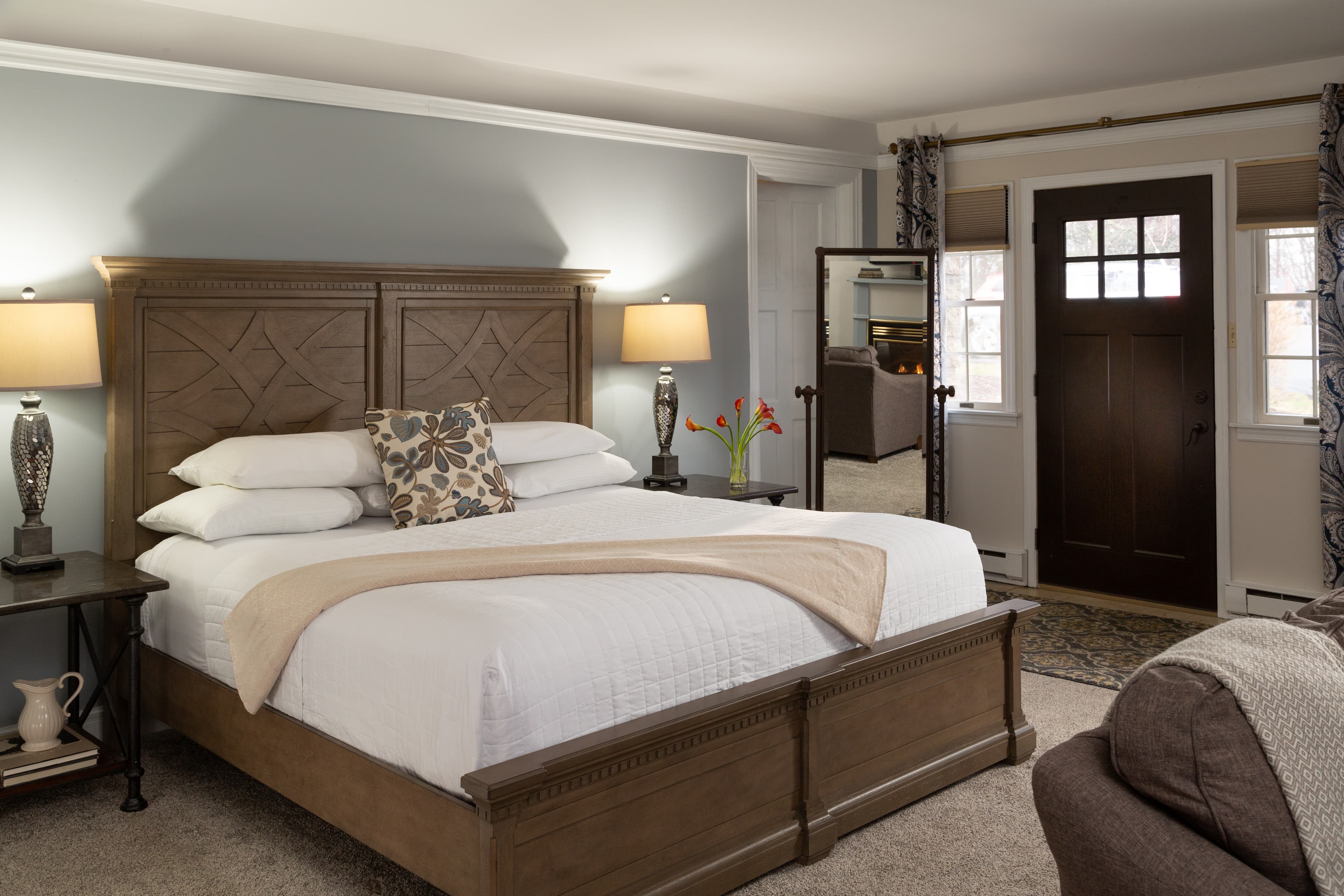 A cozy bedroom featuring a wooden bed frame, decorative pillows, and a mirror with natural light from a nearby door.