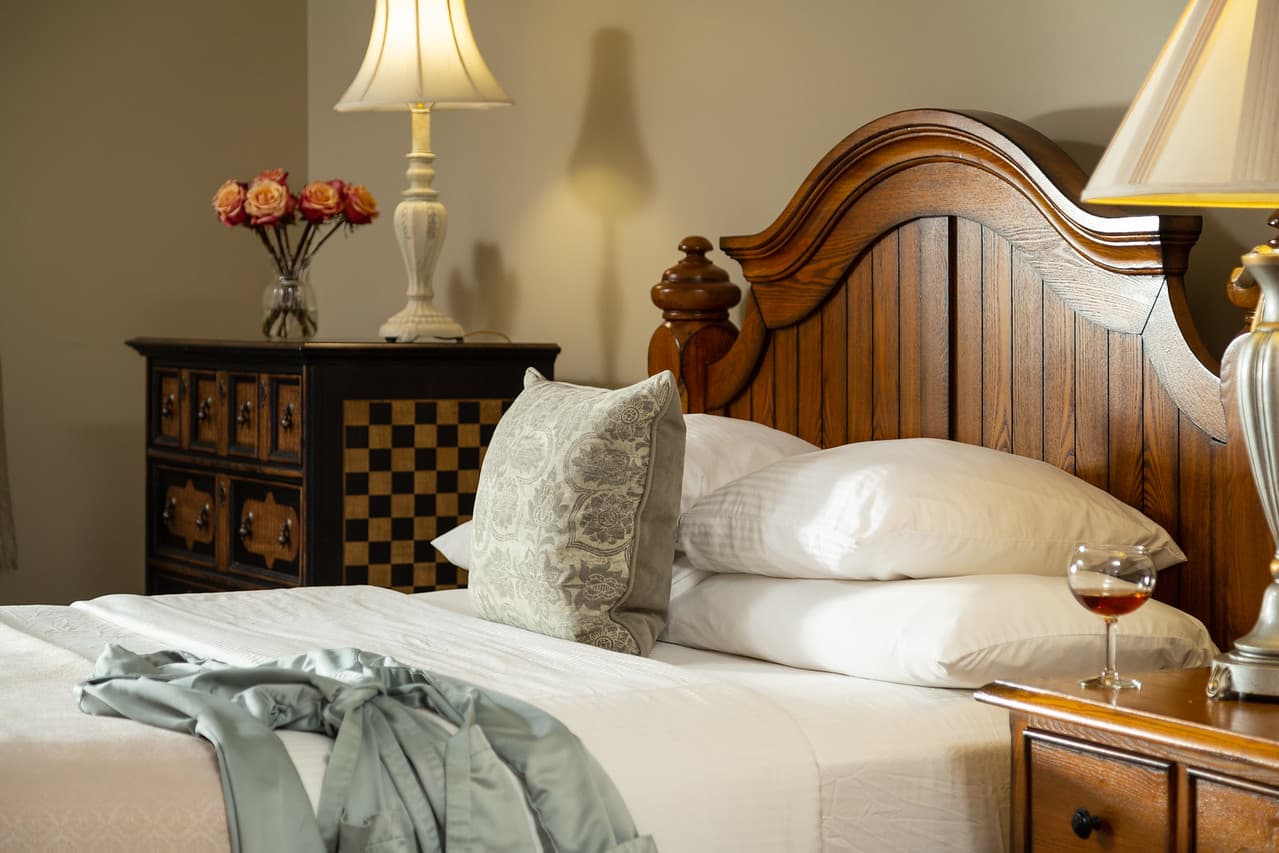 A cozy bedroom featuring a wooden bed with decorative pillows, a lamp, and a glass of wine on a bedside table.