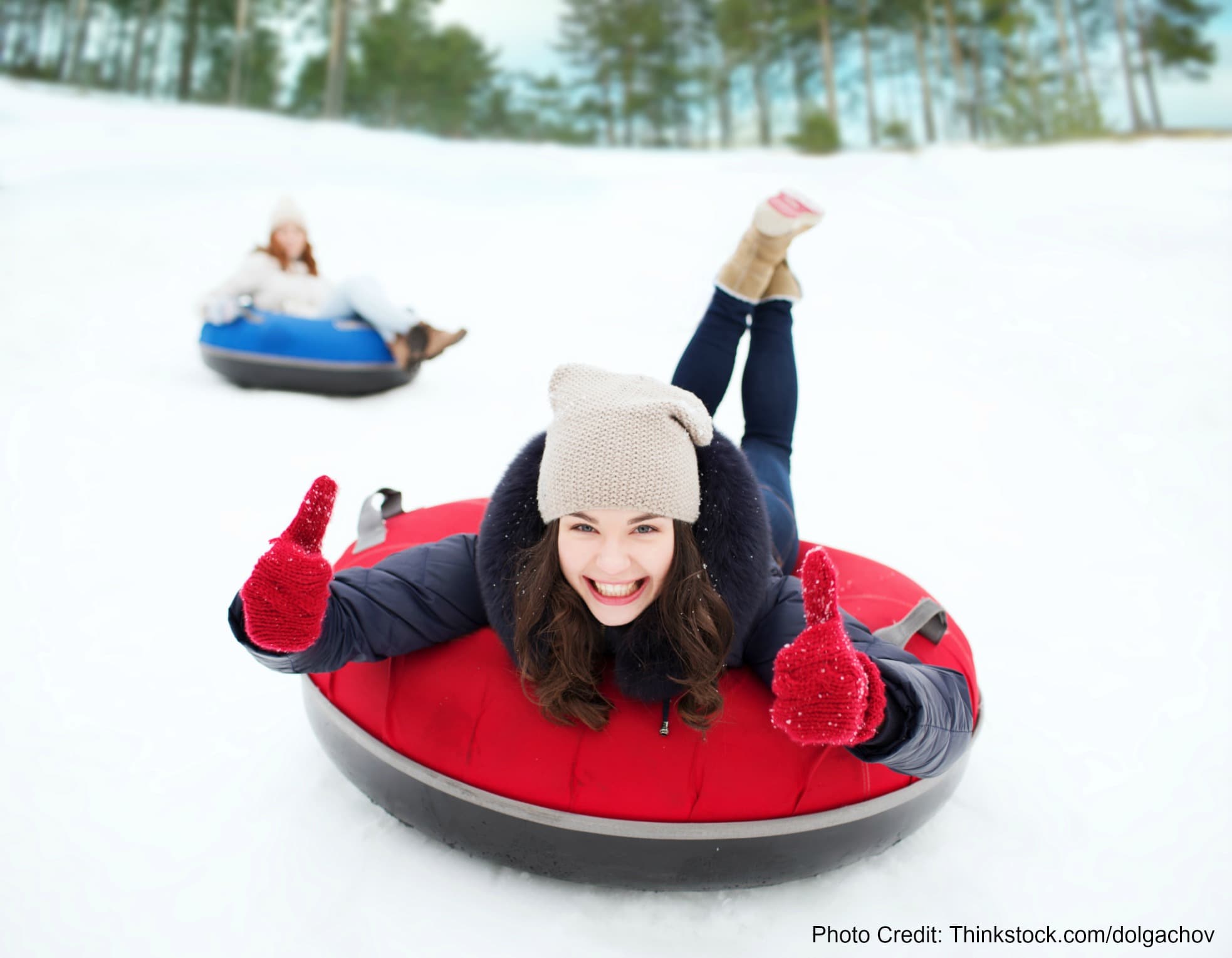 A smiling woman in winter attire gives thumbs up while sledding down a snowy hill.