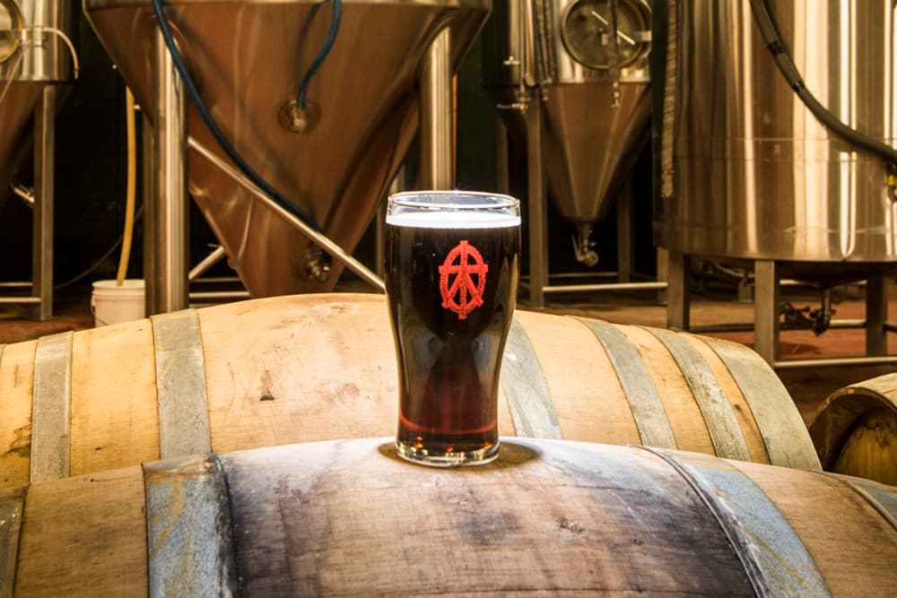 A pint glass of dark beer with the Anarchy Brewing Company logo sitting atop a wooden barrel in a brewery.