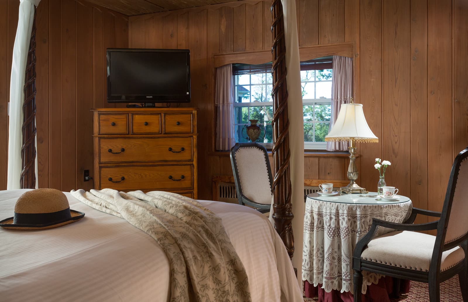 Cozy bedroom with wooden walls featuring a four-poster bed, a dresser, a small table, and a TV.