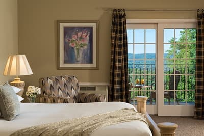 A cozy hotel room featuring a bed, patterned armchair, lamp, and large balcony doors with scenic views.
