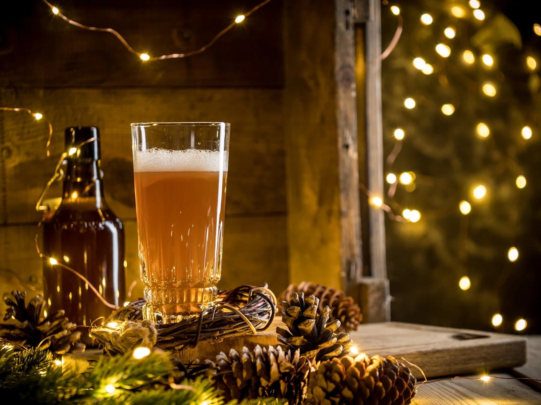 A glass of beer and a bottle are surrounded by pinecones and fairy lights.