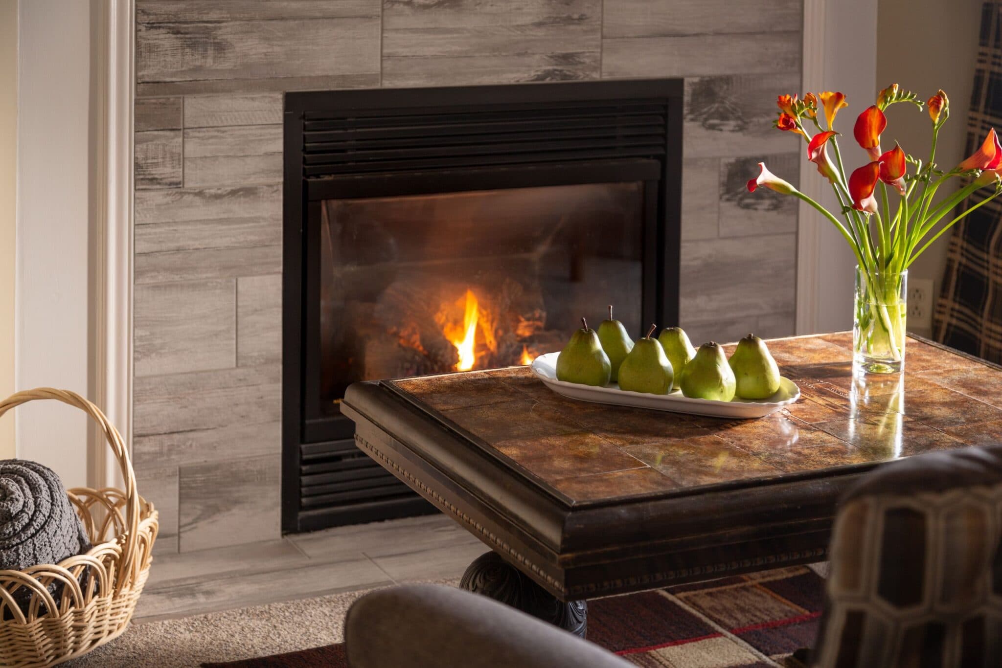 A cozy living room scene featuring a fireplace, a wooden table with green pears, and a vase of flowers.