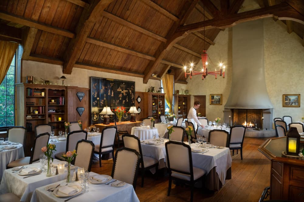 A cozy and elegant dining room with a vaulted ceiling and exposed wooden beams. Tables with white tablecloths and chairs are arranged throughout the room, and a fireplace is located in one corner. A chandelier hangs from the ceiling, and there are bookshelves and artwork on the walls. A waiter is seen setting a table.