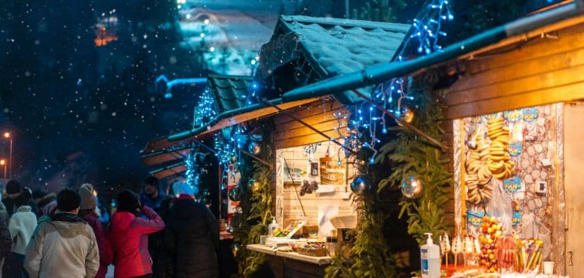 A bustling winter market scene with decorated stalls and snow falling.