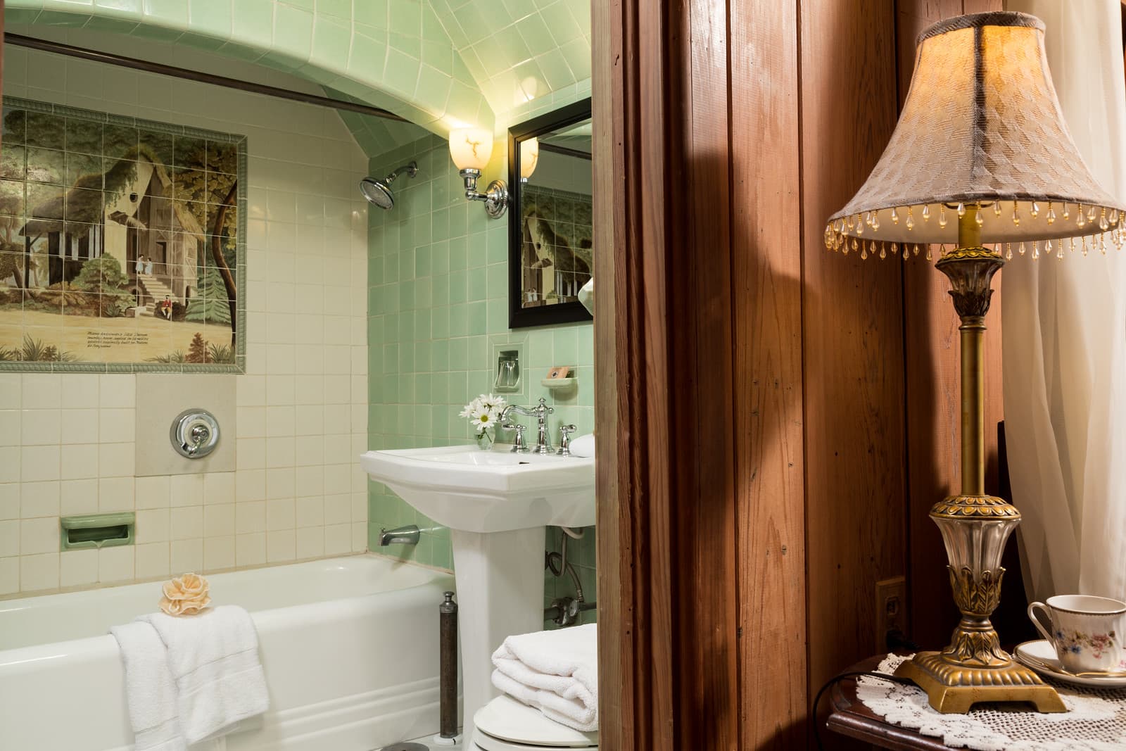 A vintage-inspired bathroom with green tiles, a tub, and a decorative lamp in the adjoining room.