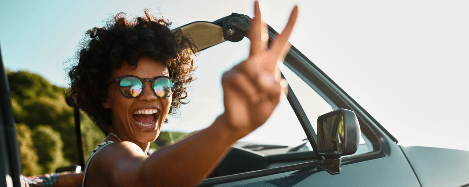 A smiling person in sunglasses flashes a peace sign from a car window. A smiling person in sunglasses flashes a peace sign from a car window.