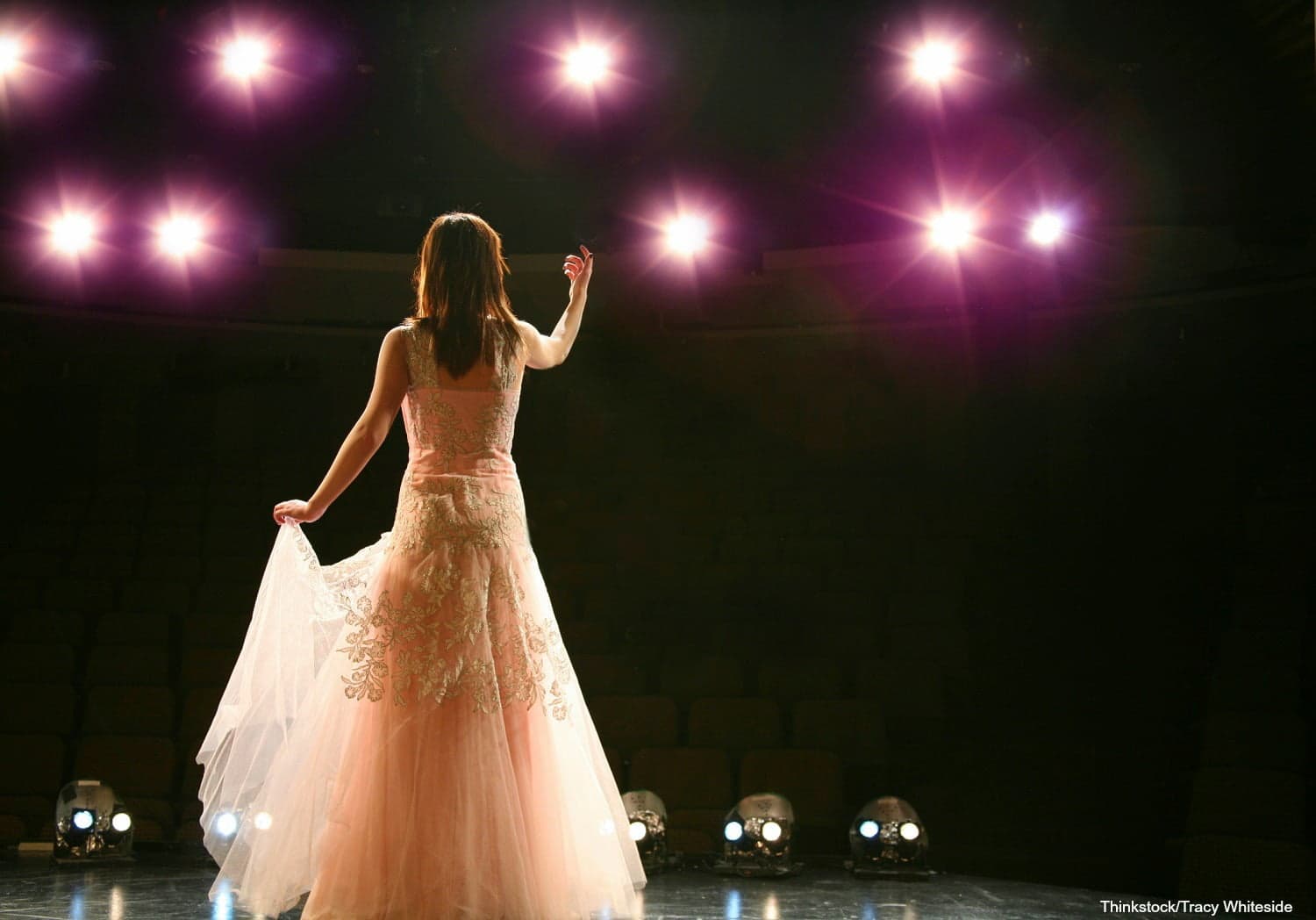A performer in a pink gown stands on stage, illuminated by bright overhead lights, with her back to the audience.