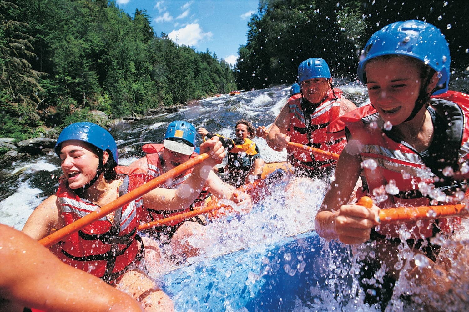 A group of people in helmets and life jackets paddles a raft through white water rapids.