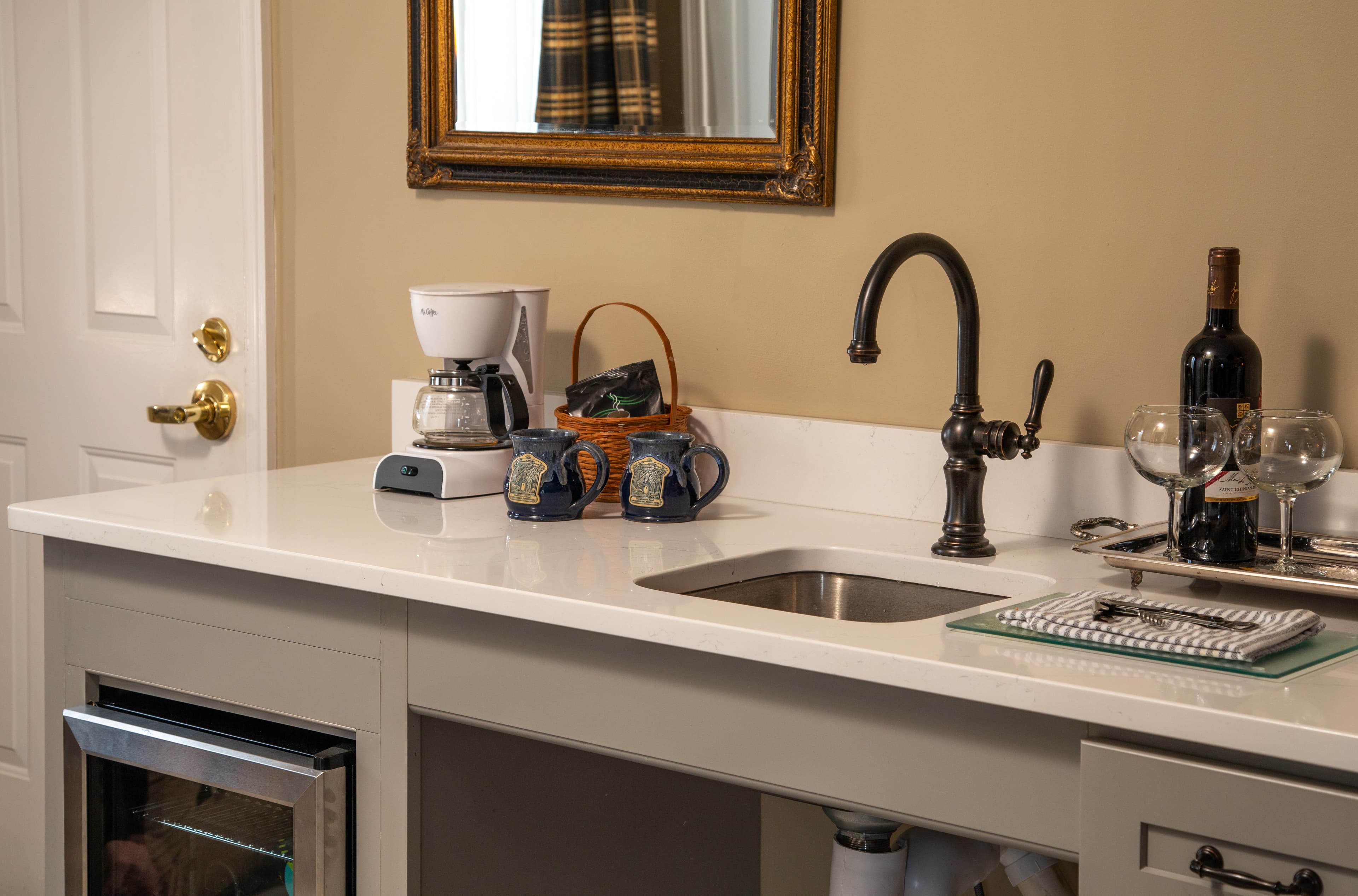 A modern kitchen counter featuring a coffee maker, wine bottles, glasses, and a sink.
