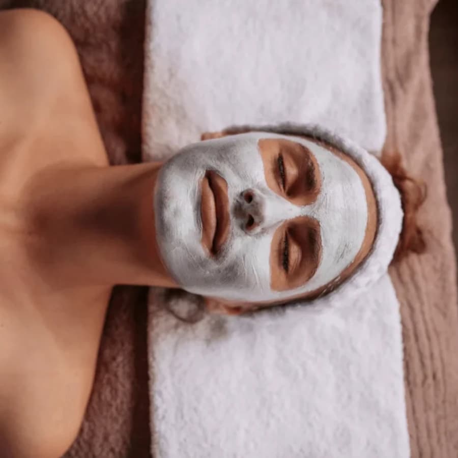 A woman lying on a table with her head on a white towel, and her face covered in a white spa mask, getting a facial