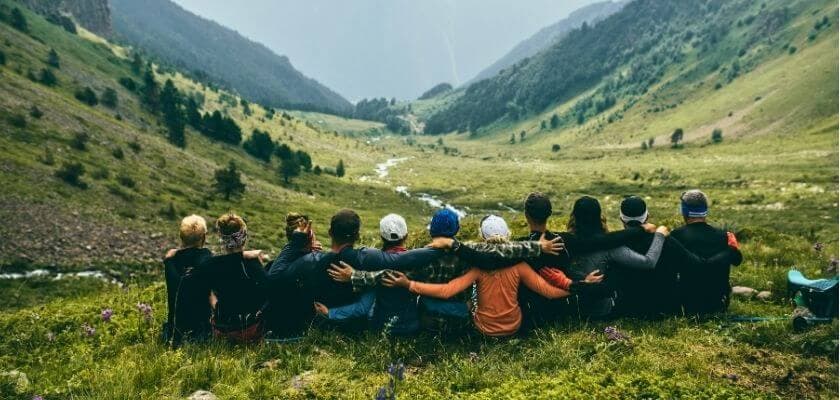 A group of people sitting together on a grassy hillside, overlooking a valley with a river and mountains in the background.