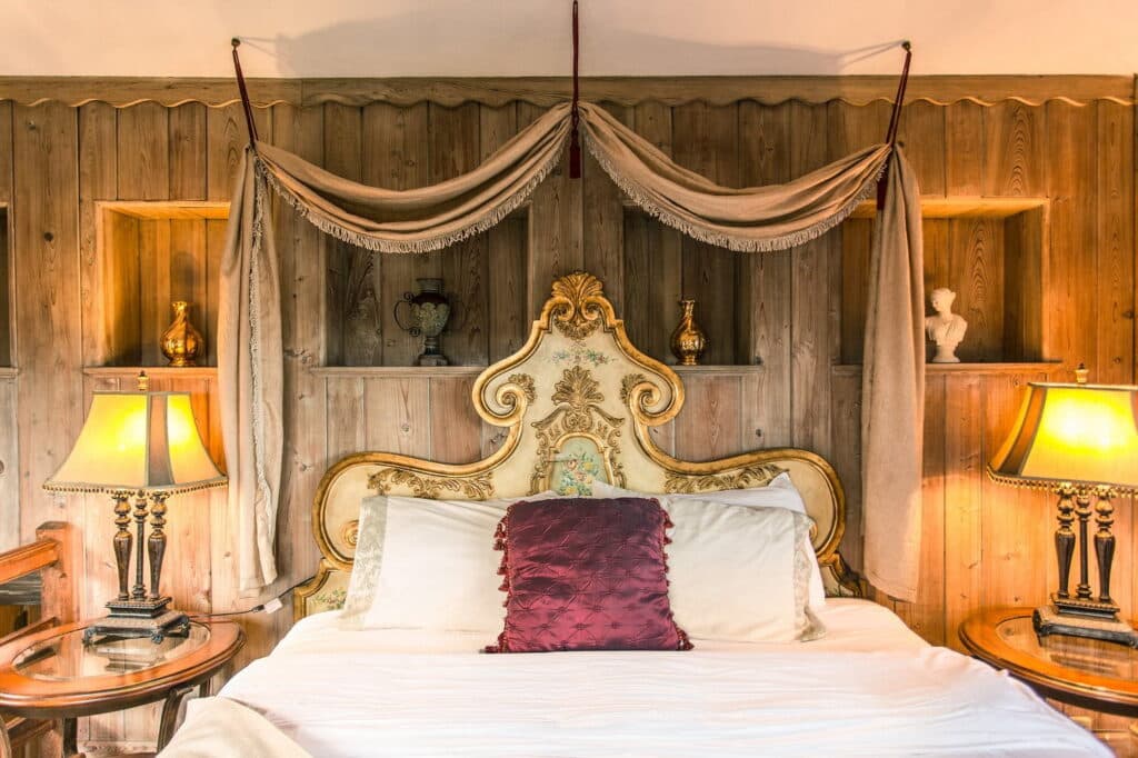 A cozy bedroom featuring an ornate headboard, plush pillows, and decorative lamps against a wooden backdrop.