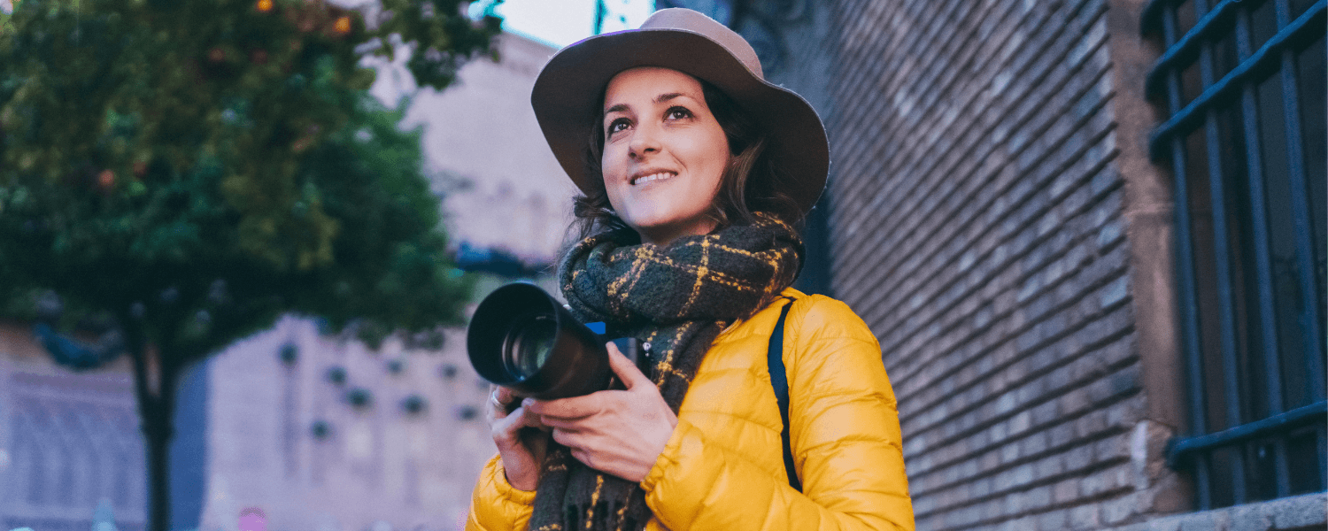 A woman wearing a yellow jacket and hat holds a camera while smiling outdoors. A woman wearing a yellow jacket and hat holds a camera while smiling outdoors.