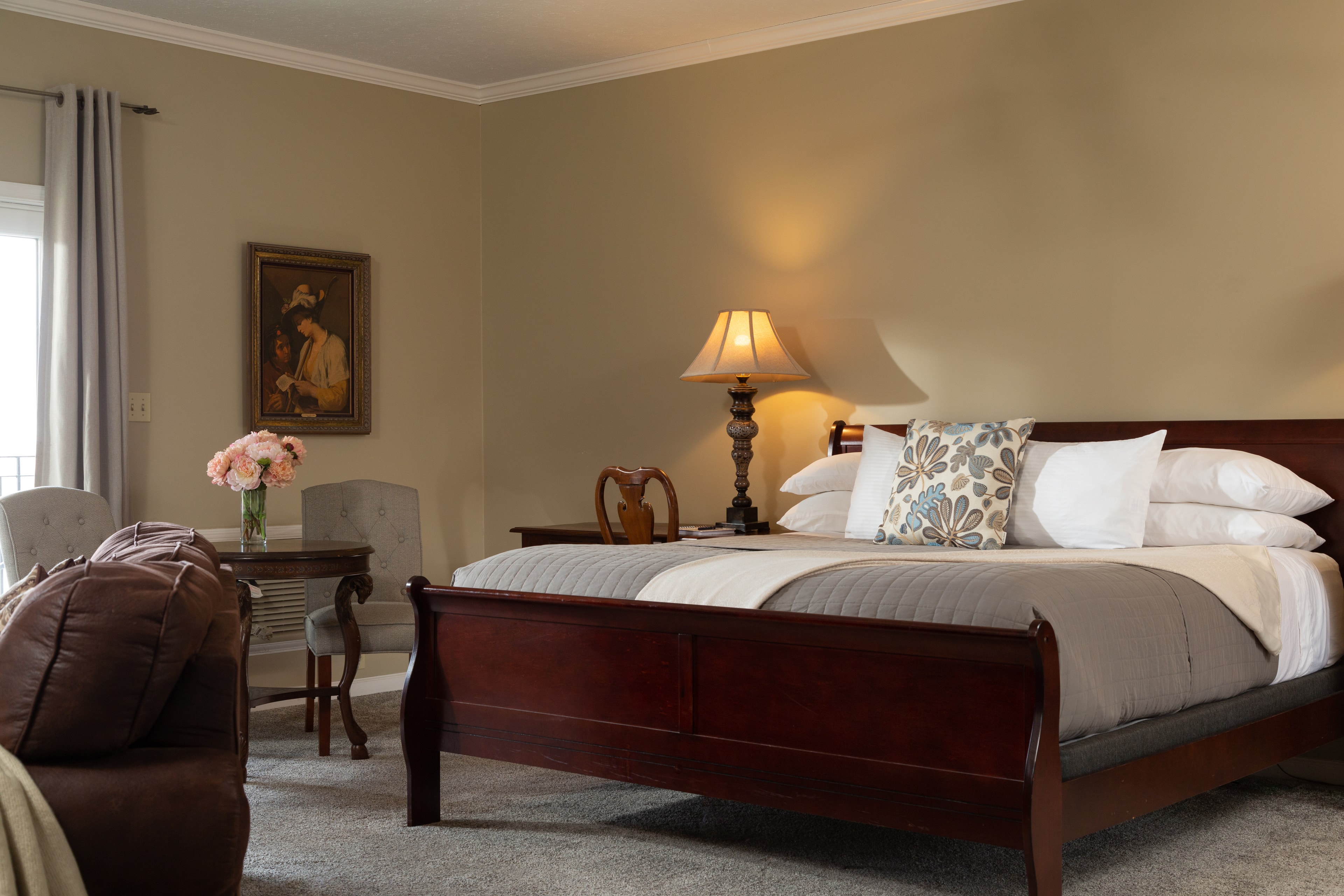 A cozy bedroom featuring a wooden bed, decorative pillows, a vintage lamp, and a small dining table with flowers.