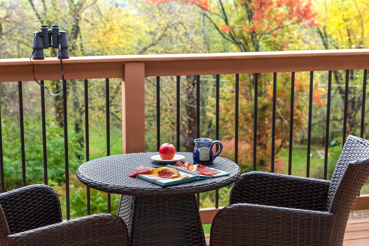 A cozy balcony featuring a table with an apple, a plate of snacks, and a coffee mug, surrounded by colorful autumn foliage.