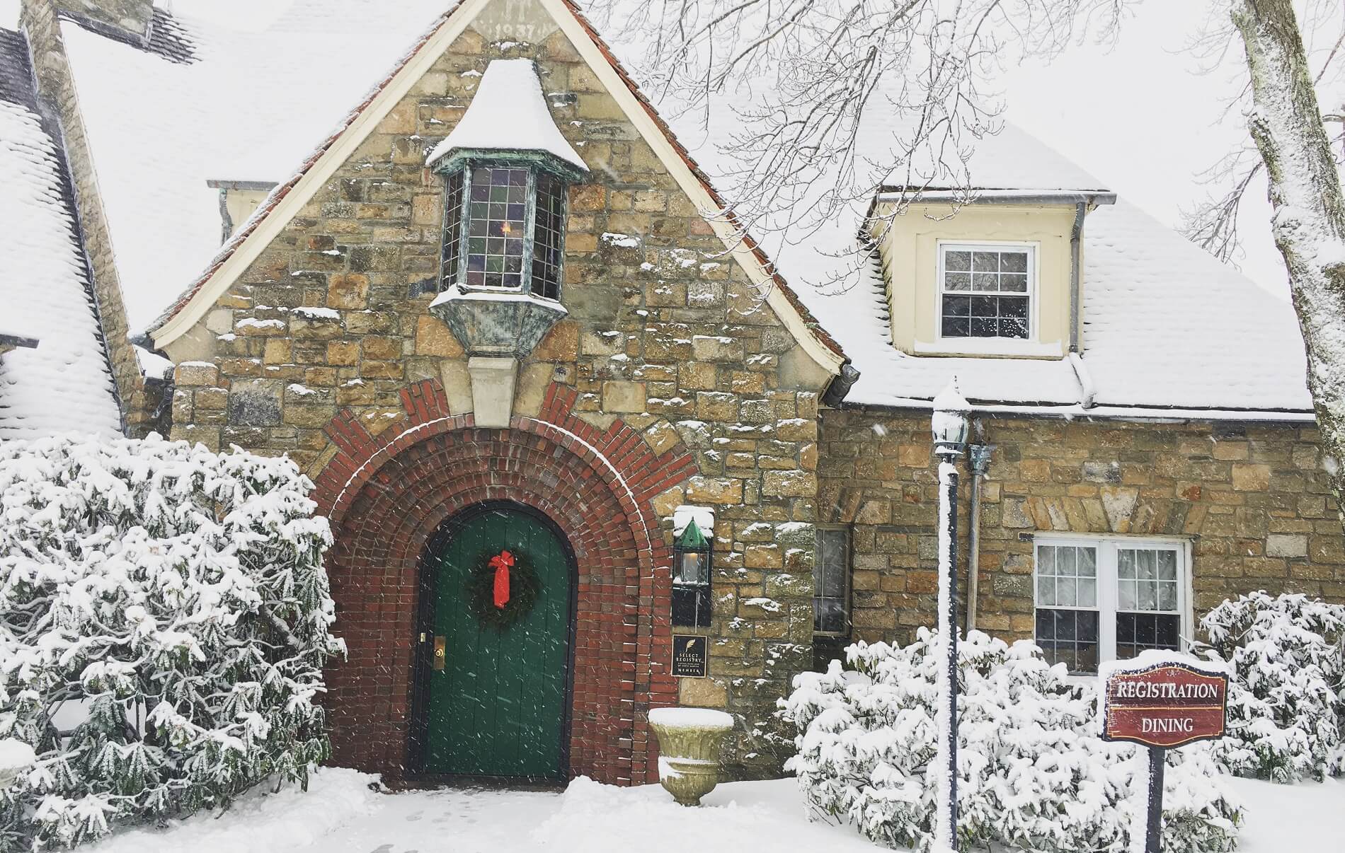 A stone house adorned with a festive wreath, surrounded by snow-covered trees and a sign for registration and dining.