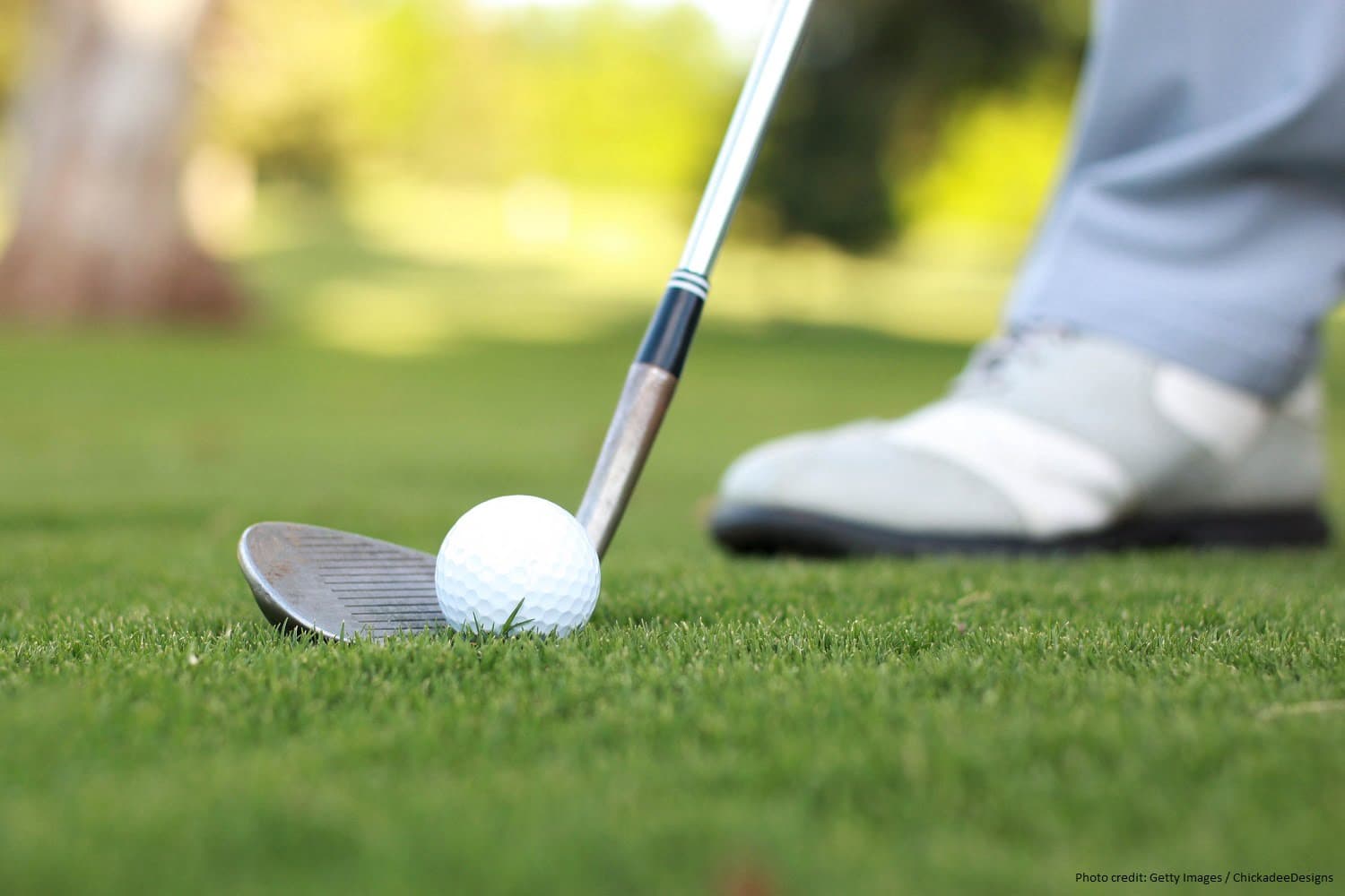 A golf club is poised just above a white golf ball on a grassy course.