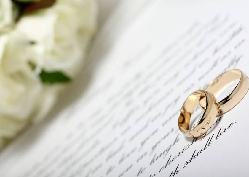 Two gold wedding rings resting on a wedding vow document beside a bouquet of flowers. Two gold wedding rings resting on a wedding vow document beside a bouquet of flowers.