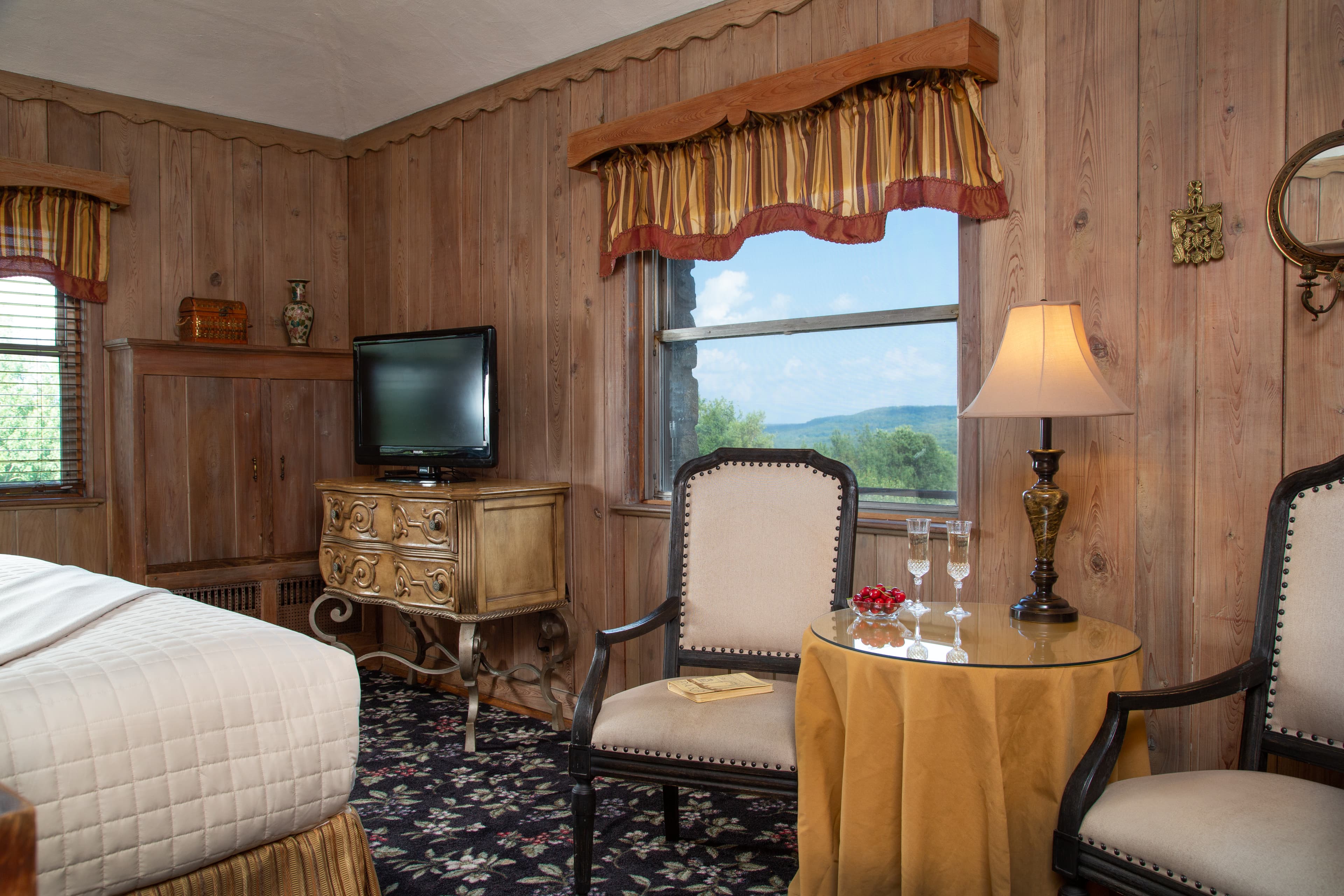 A cozy hotel room featuring wood paneling, a TV, two chairs, and a side table with glasses and decorative items.