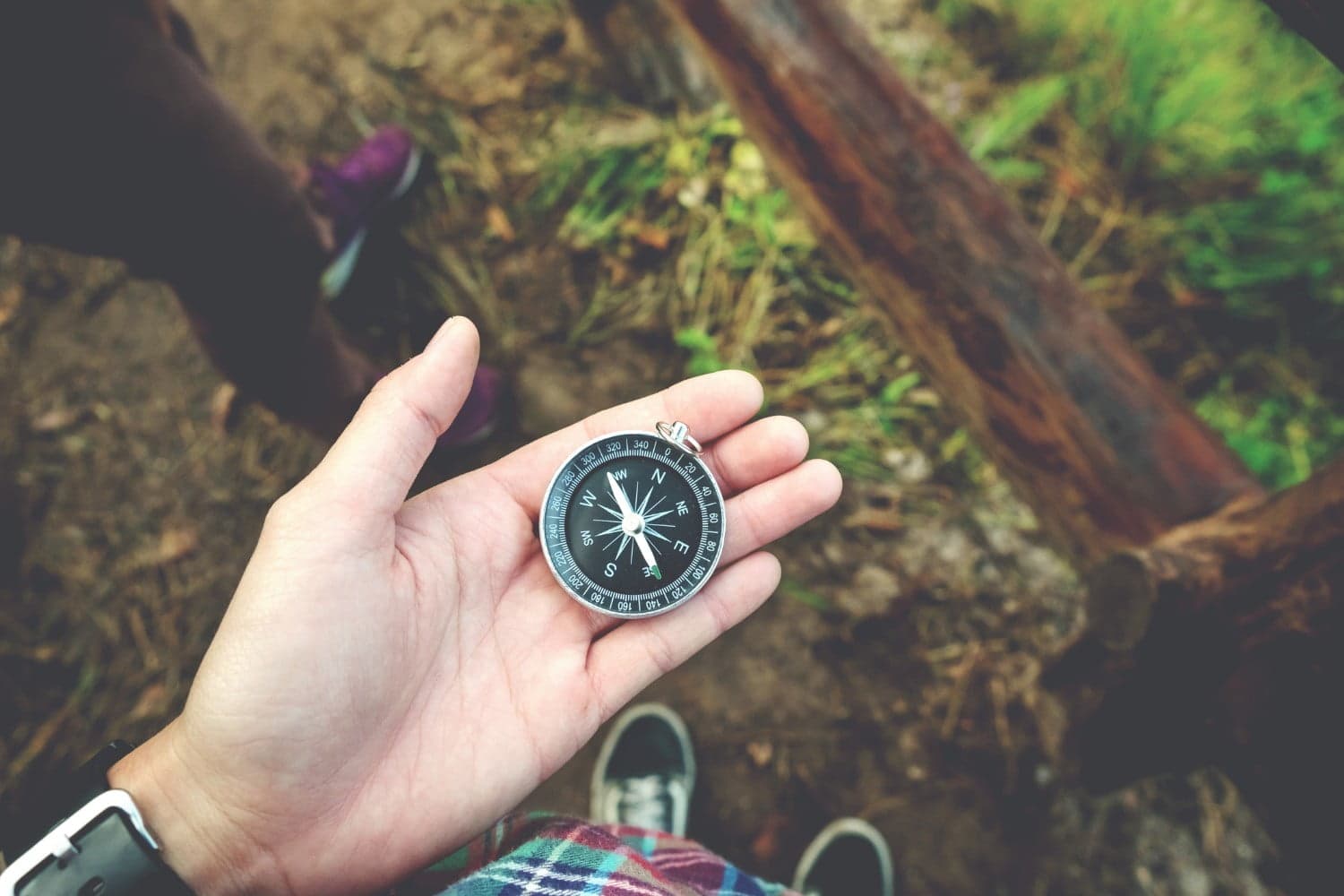 A hand holding a silver compass with a black face, set against a blurred natural background.