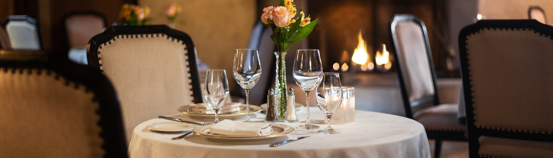 A beautifully set table in a cozy restaurant. A white tablecloth covers the round table, and there are wine glasses, plates, and silverware arranged for dining. A vase of flowers sits in the center, and a fireplace is visible in the background.