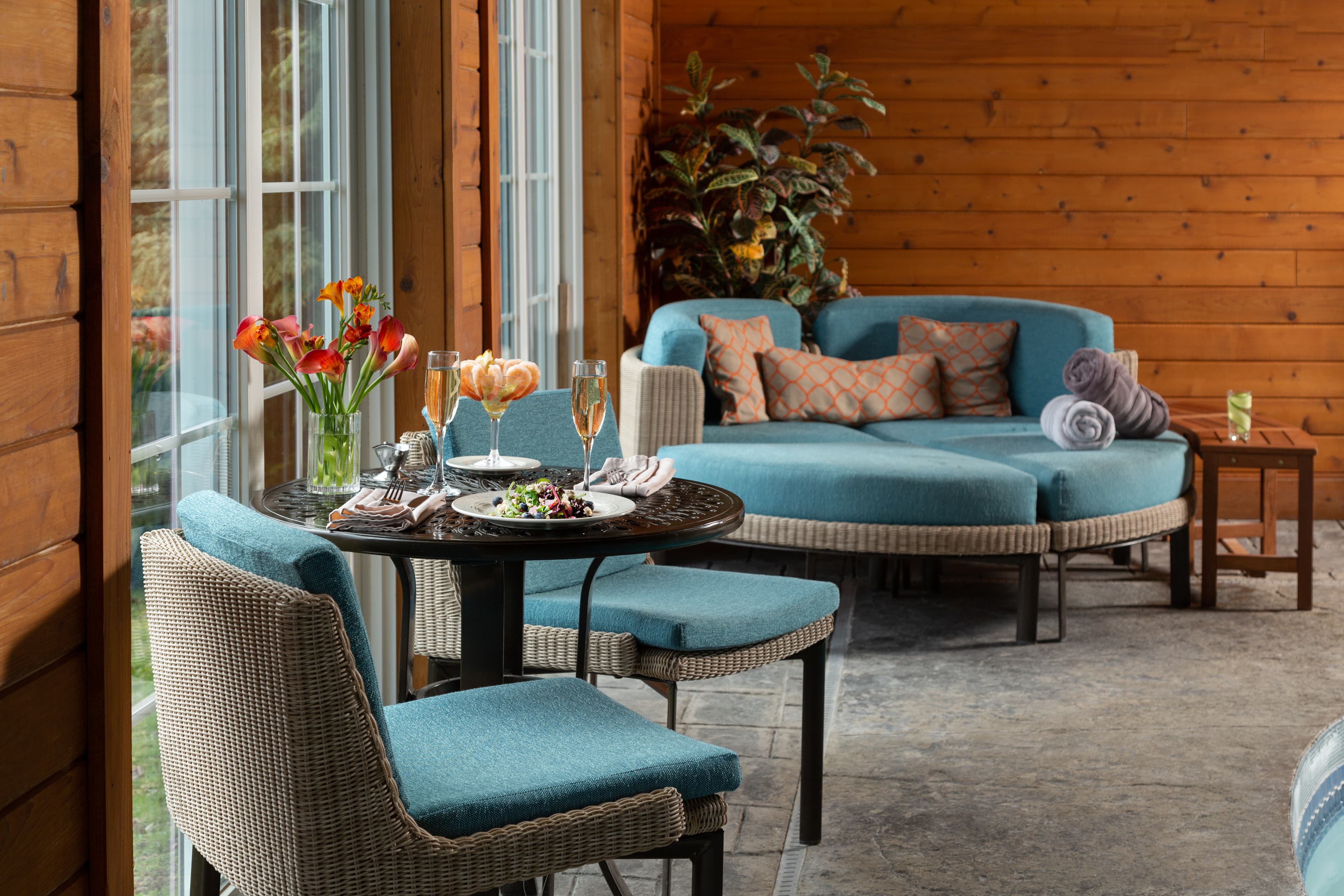 A cozy indoor seating area with large windows overlooking a lush green landscape. A round table with chairs sits next to a curved sofa with pillows. There are flowers and champagne glasses on the table, and a small table with towels and a drink is nearby. The walls are paneled with wood, and there is a potted plant in the corner.
