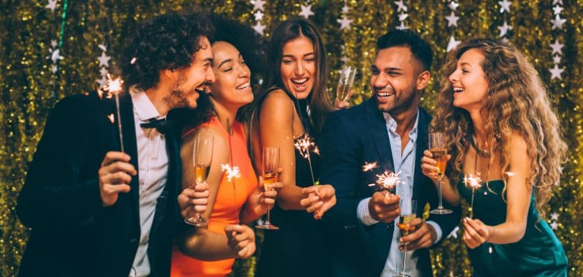 A group of five friends celebrate together with drinks and sparklers against a festive backdrop.