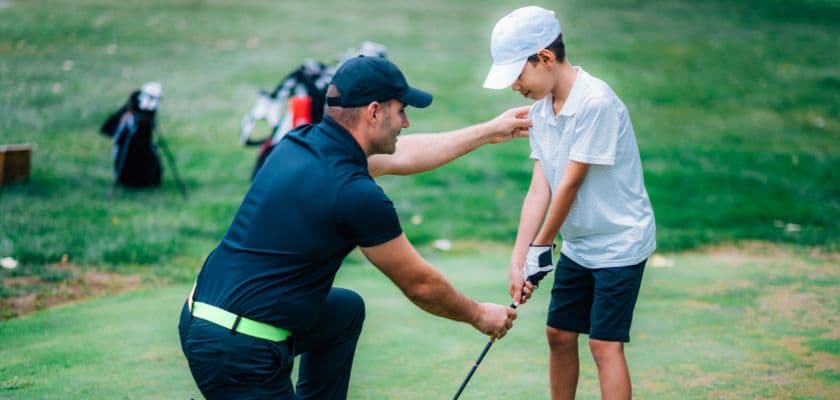 Adult helping a child practice golf on a course, guiding their stance and grip during a lesson